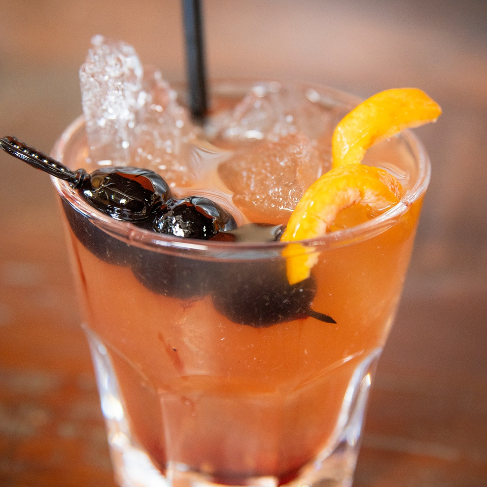 A refreshing cocktail with ice, dark cherries on a pick, and an orange peel garnish.