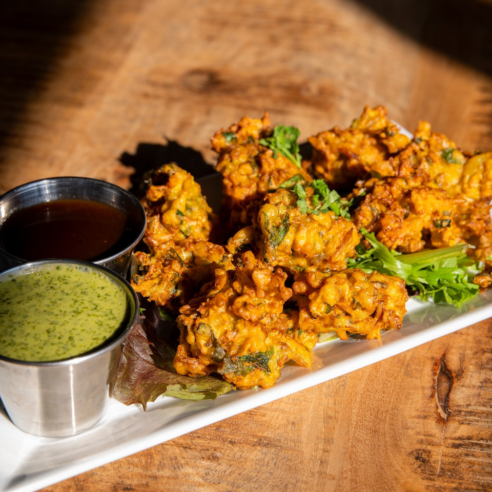 Crispy golden-brown pakoras with fresh herbs, served with two dipping sauces on a white plate.